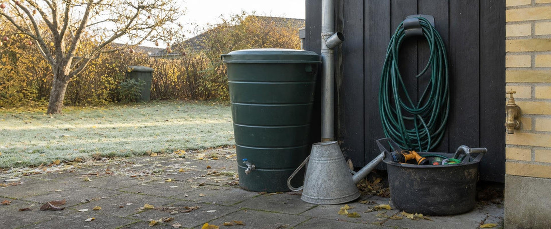 Tom regnvandstønde med åben hane, oprullet haveslange på skurvæg og omvendt zinkvandkande i en efterårshave med rimfrost
