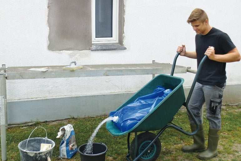 Mand tømmer vandsæk fra trillebør i spand ved husmur