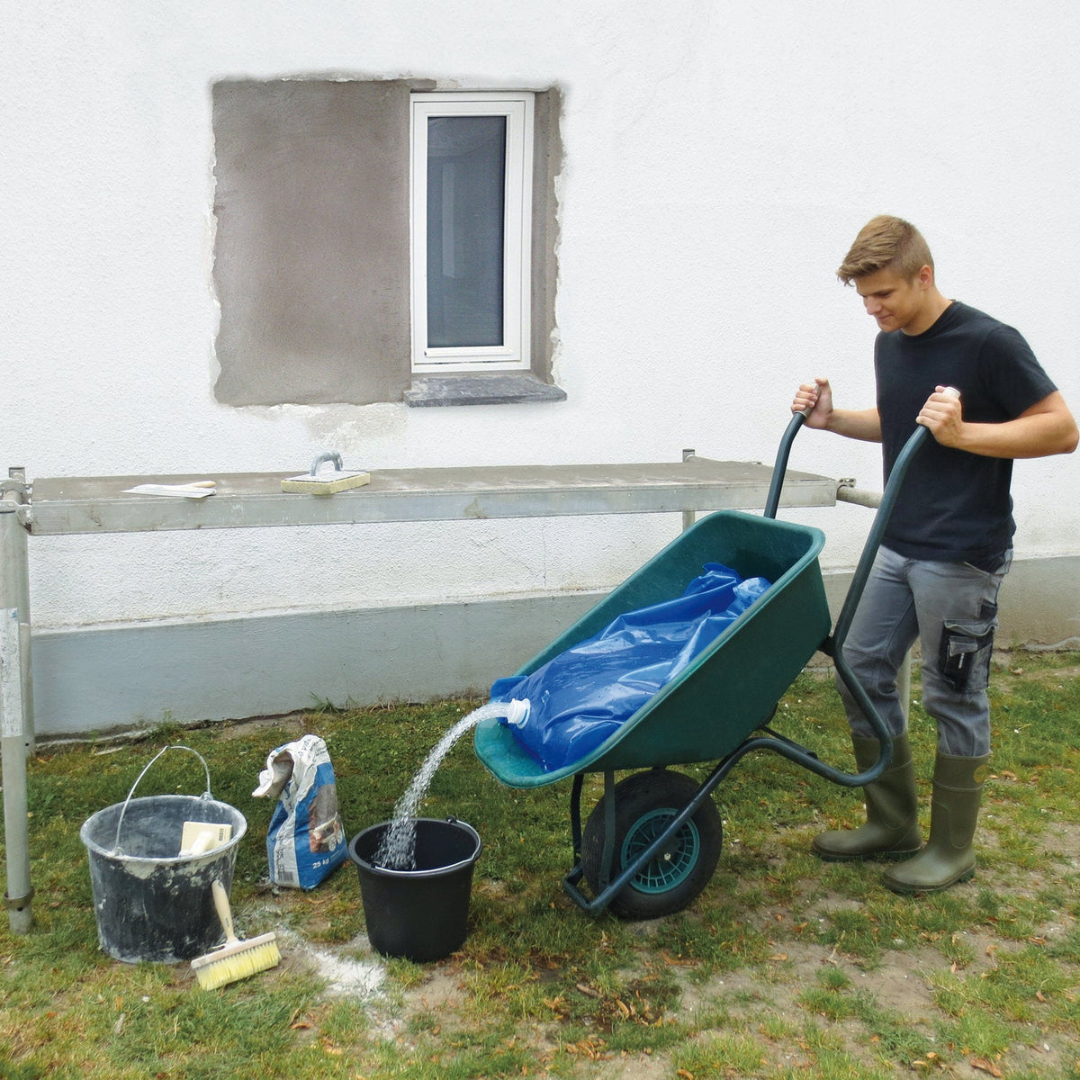 Mand tømmer vandsæk fra trillebør i spand ved husmur